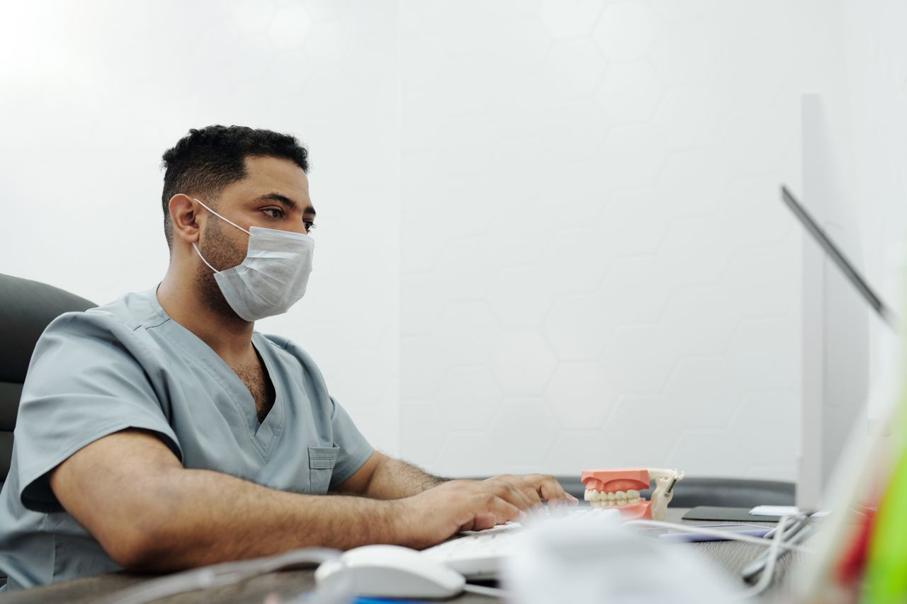 Medical professional in scrubs and mask working on computer in clinic office.